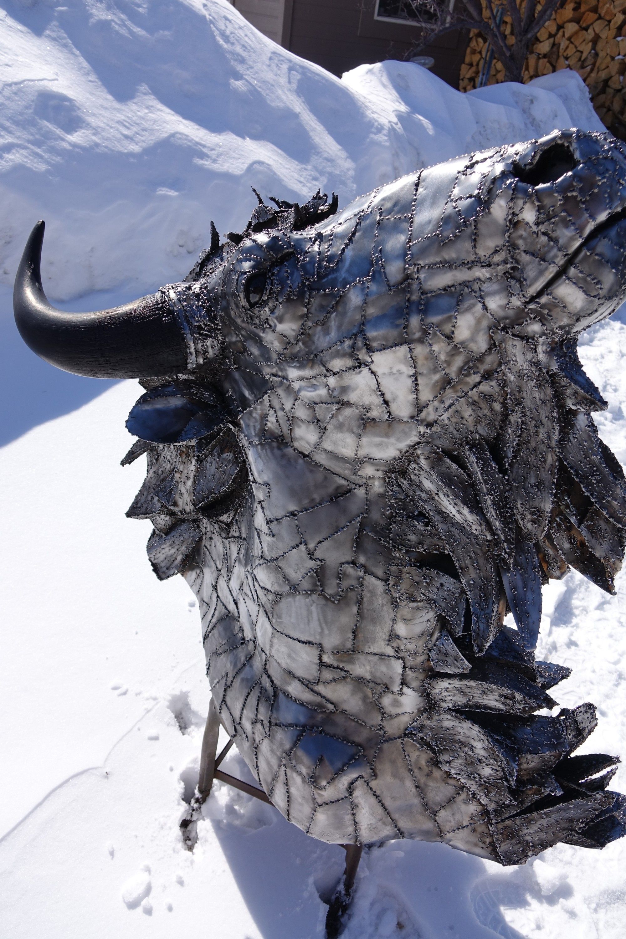 The steel bison head from the other side, its shaggy mane built up from overlapping steel plates, a woodpile behind it
