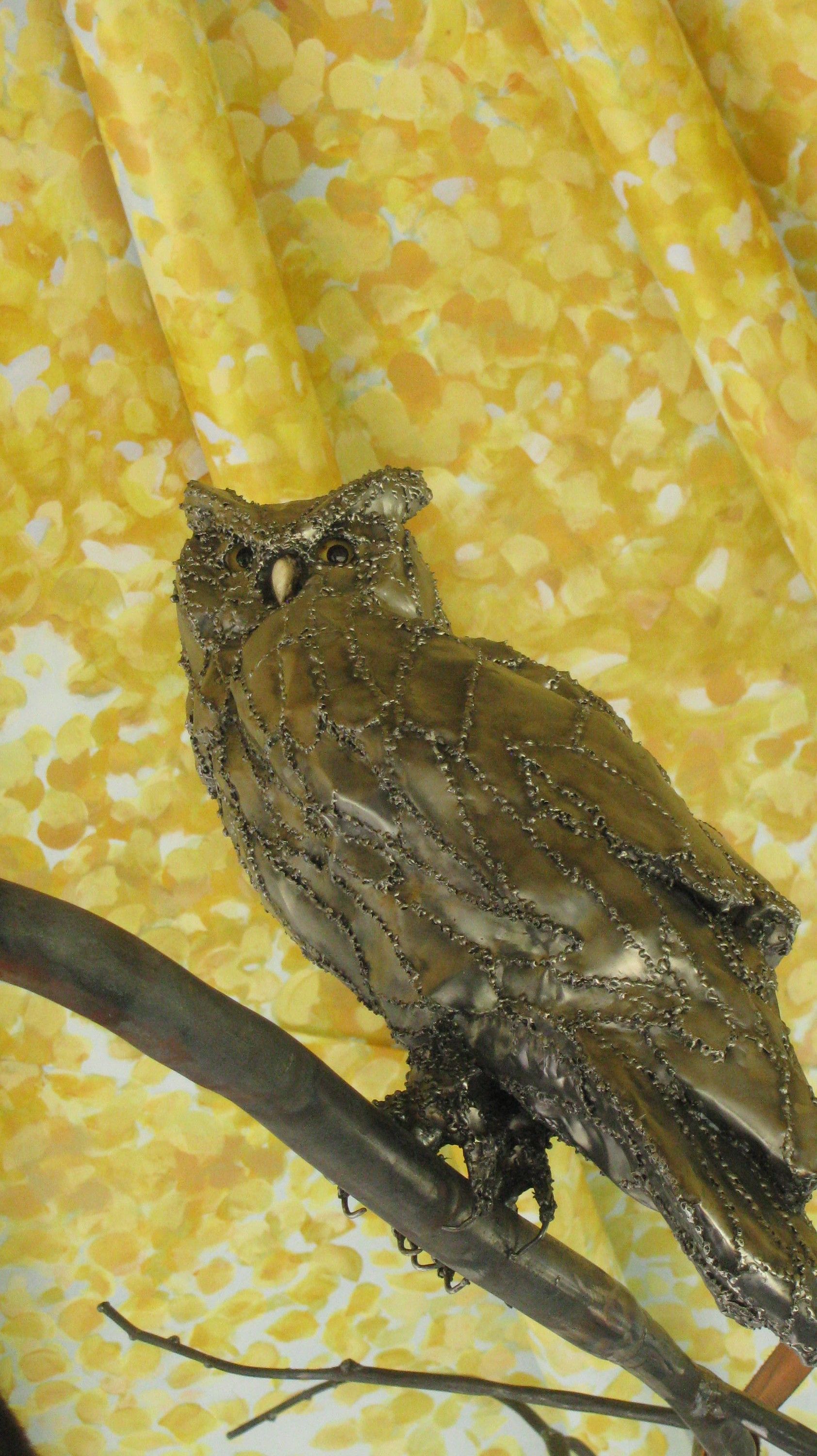 The whole steel owl perched on a steel branch, head turned toward the camera, against a painted backdrop of yellow leaves