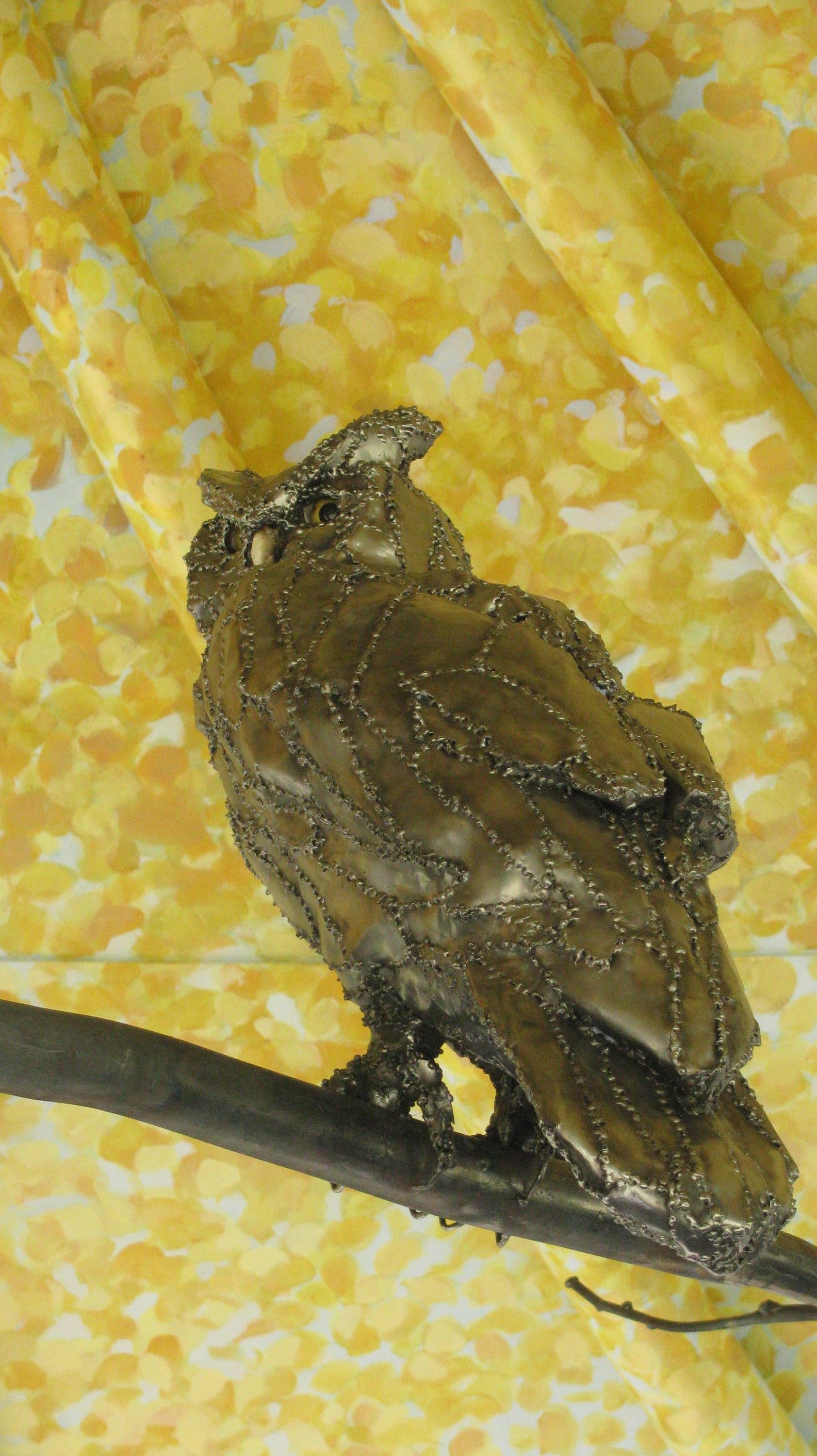 The steel owl seen from behind on its branch, showing the built-up plates across its back and folded wings