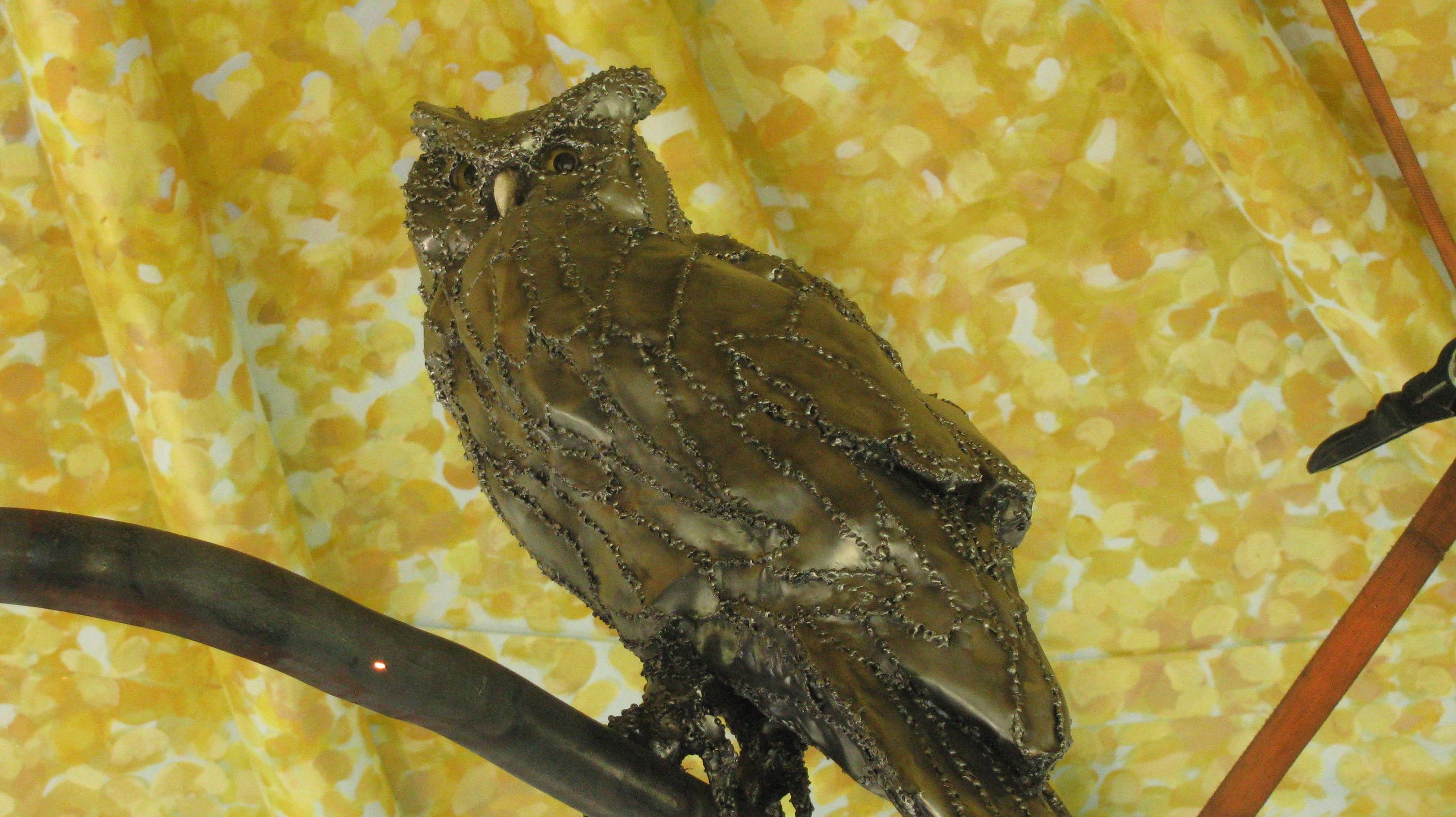 The steel owl close from the side, the welded seams between its steel feather plates catching the light