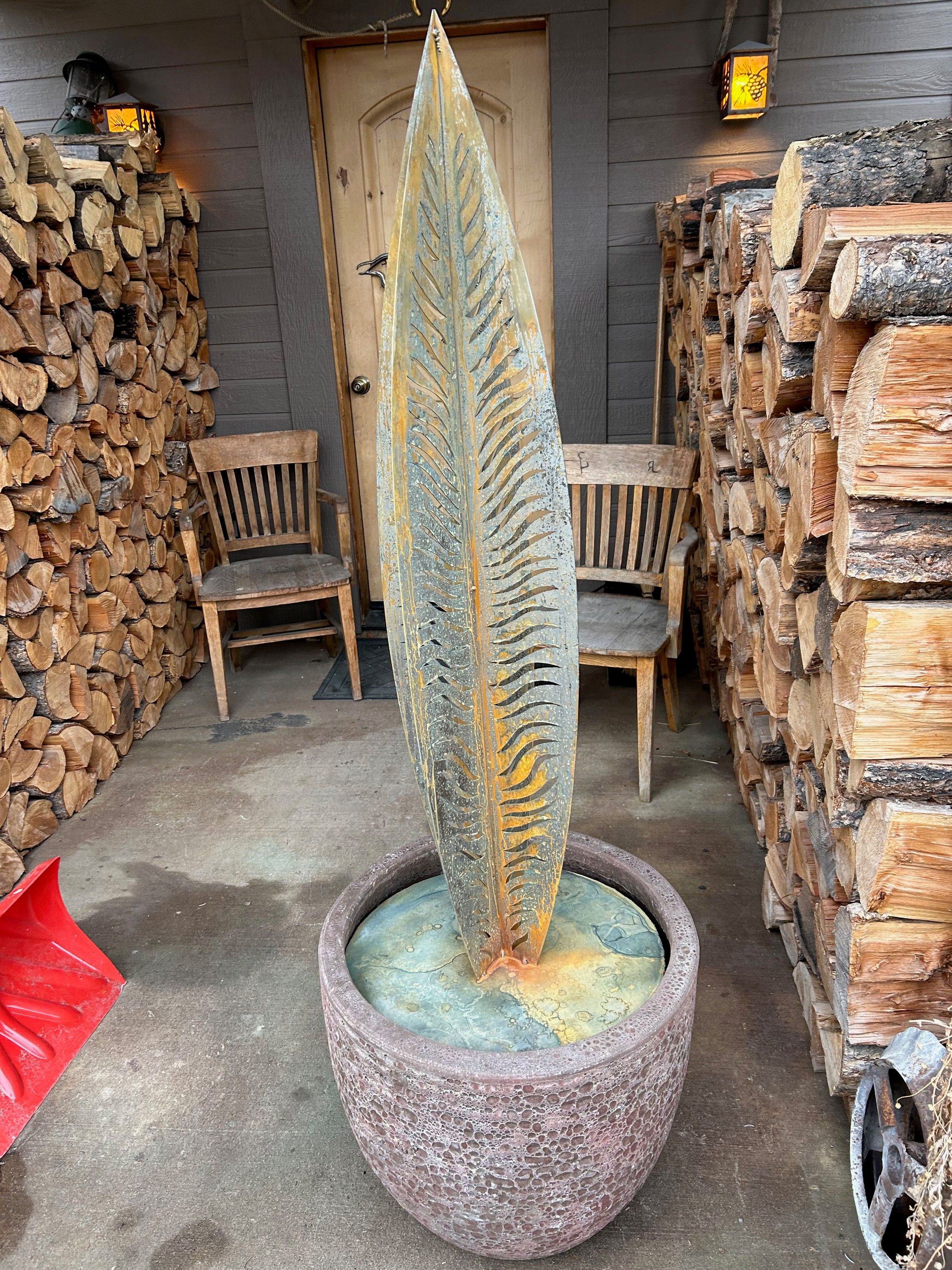 A tall steel silver fern frond standing upright in a stone planter on a covered porch, its cut leaflets catching the light, stacked firewood on either side