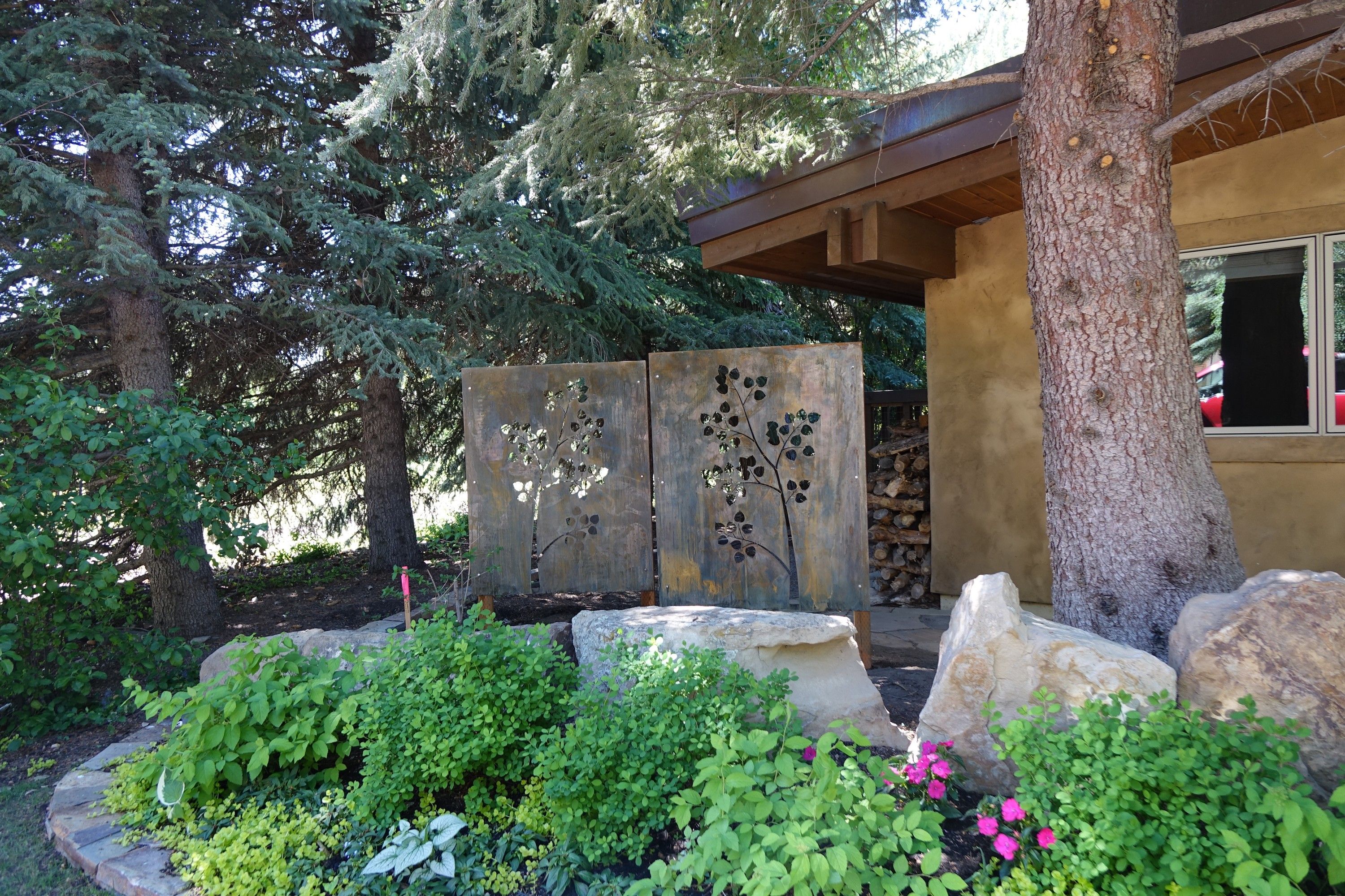 Two patina steel panels cut with aspen branches and leaves, standing on wooden feet in a planting bed beside a house