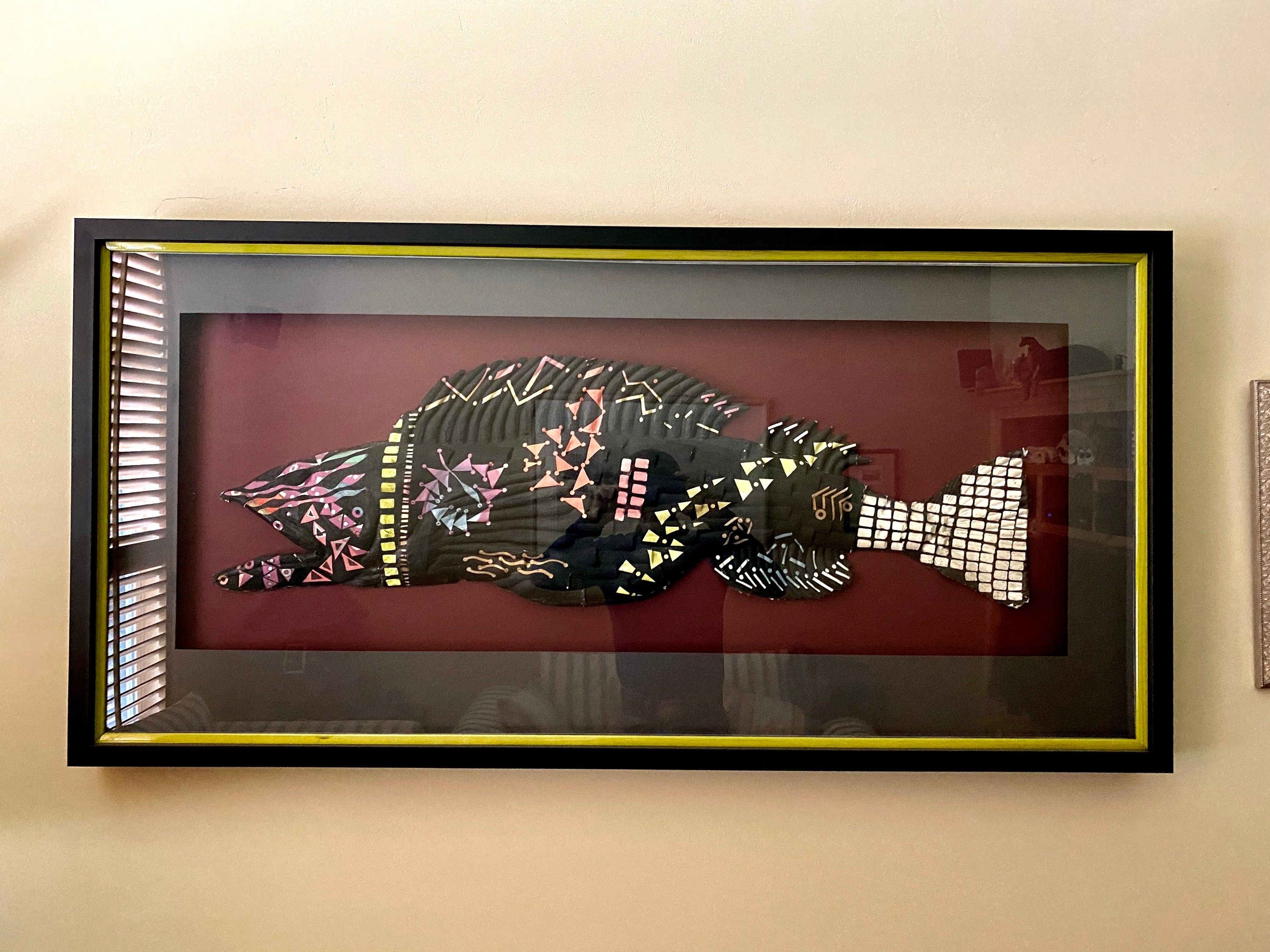 A stylised ling cod in paper mache, its dark body patterned with pink, yellow and white geometric marks and a blocky white tail, mounted on a deep red ground in a black and gold shadow box