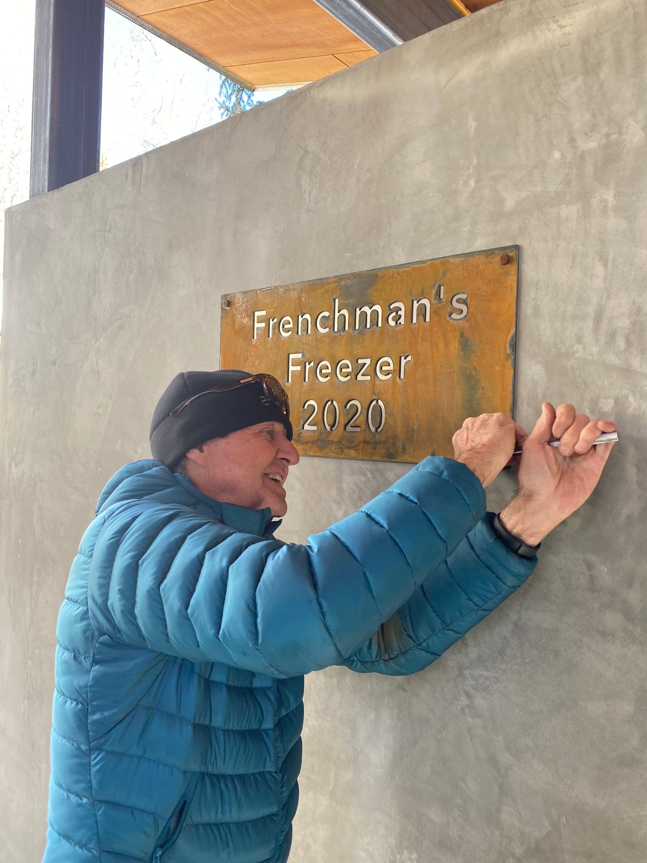 A patinated steel plaque with polished stainless letters reading Frenchman's Freezer 2020, with Russ Lamb fastening it to a concrete wall