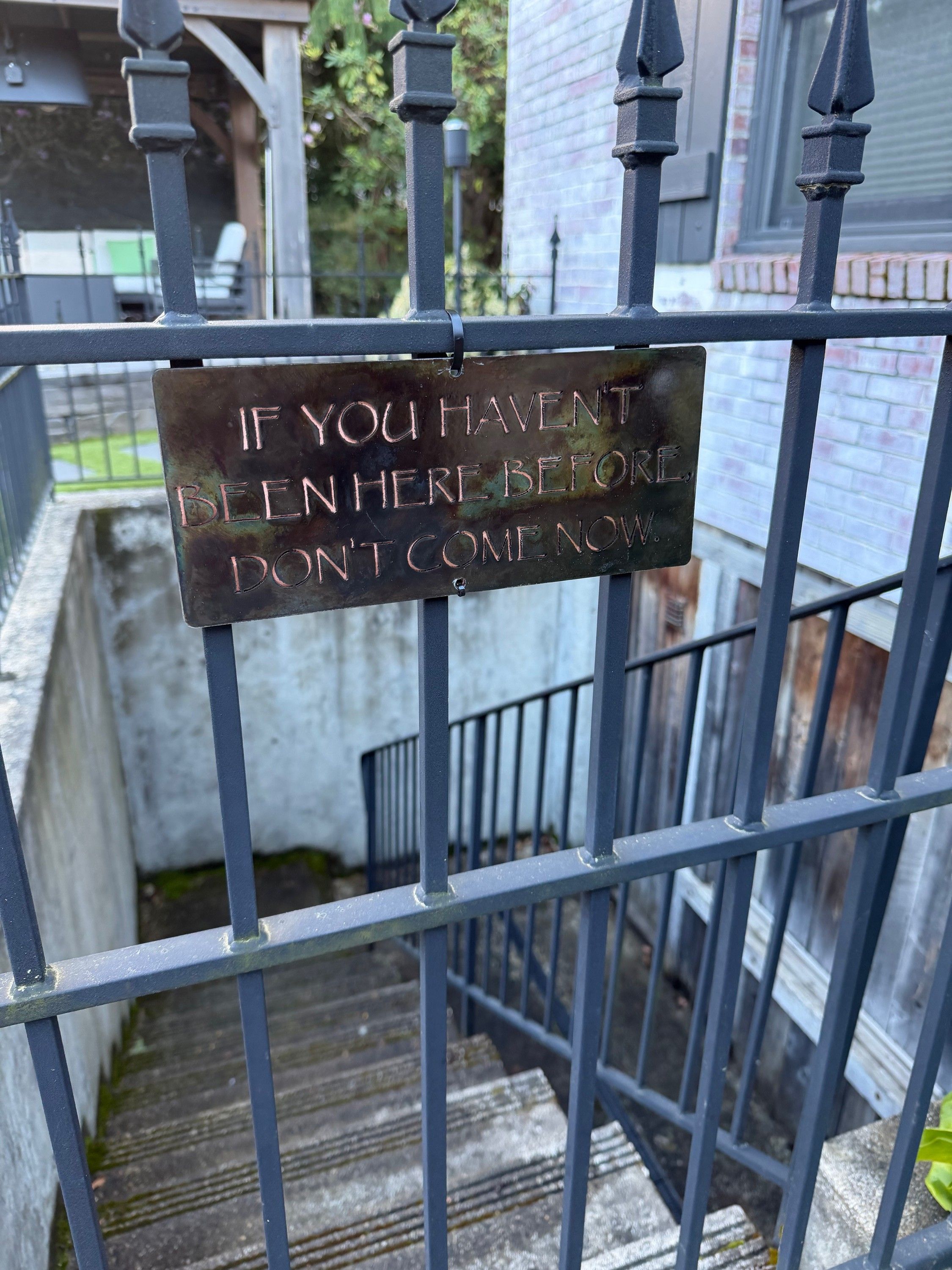A laser cut steel sign hanging on an iron gate, its raised letters reading IF YOU HAVENT BEEN HERE BEFORE DON'T COME NOW, with steps descending behind