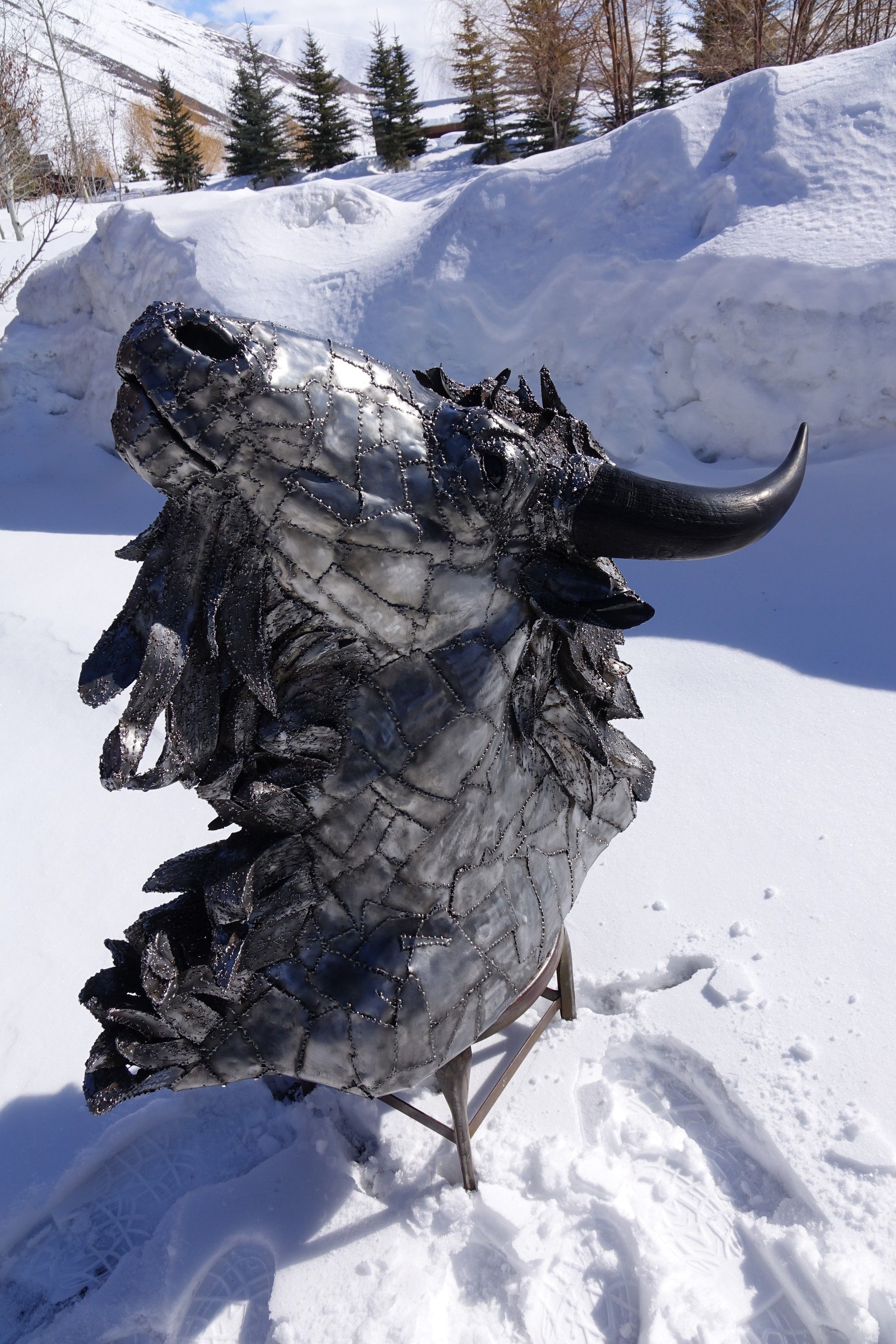A steel bison head on a stand in deep snow, muzzle raised, one dark horn curving up against a snowbank and conifers