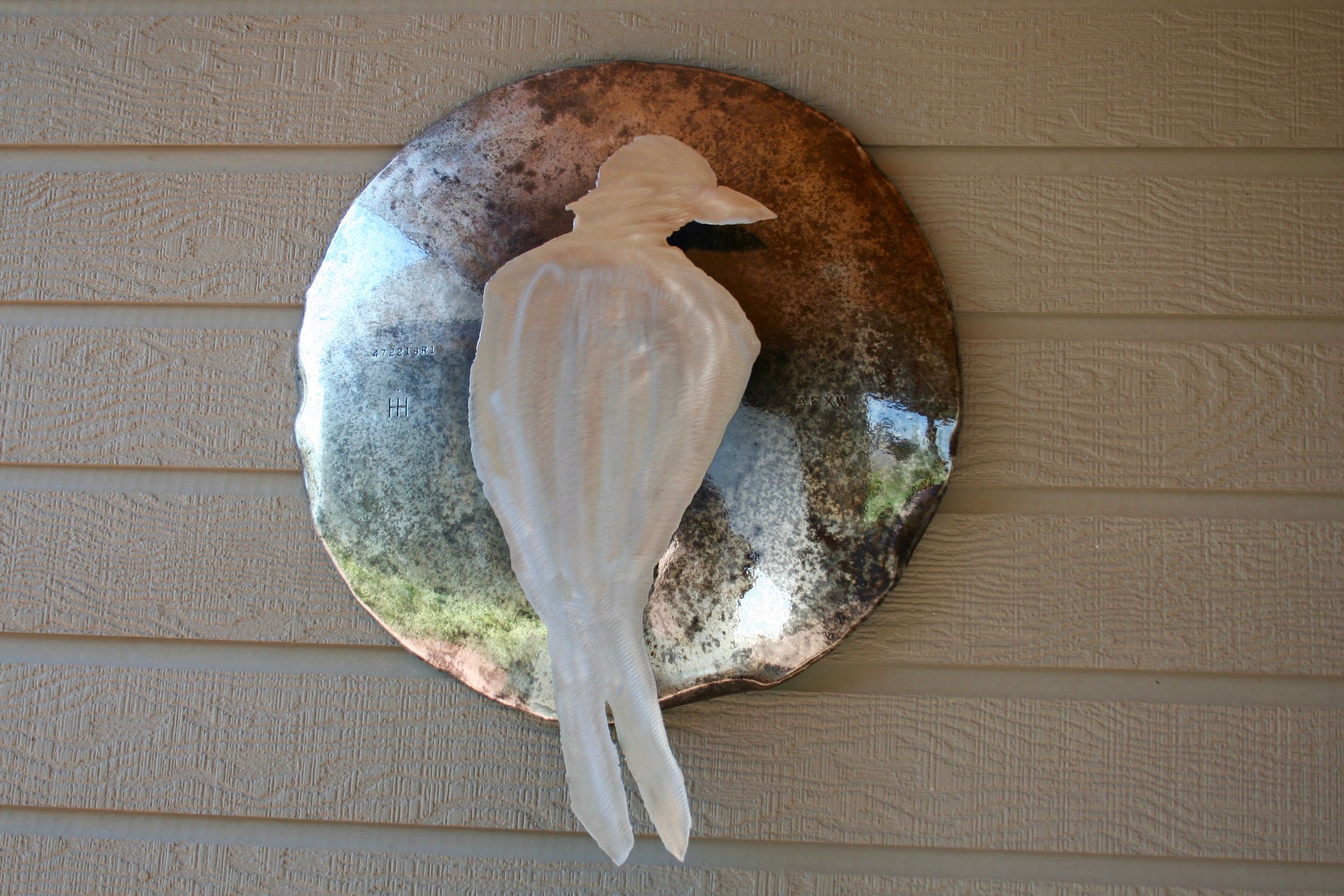 A pale steel crow standing against a weathered hubcap that reads as a full moon, hung on the lap siding of a house