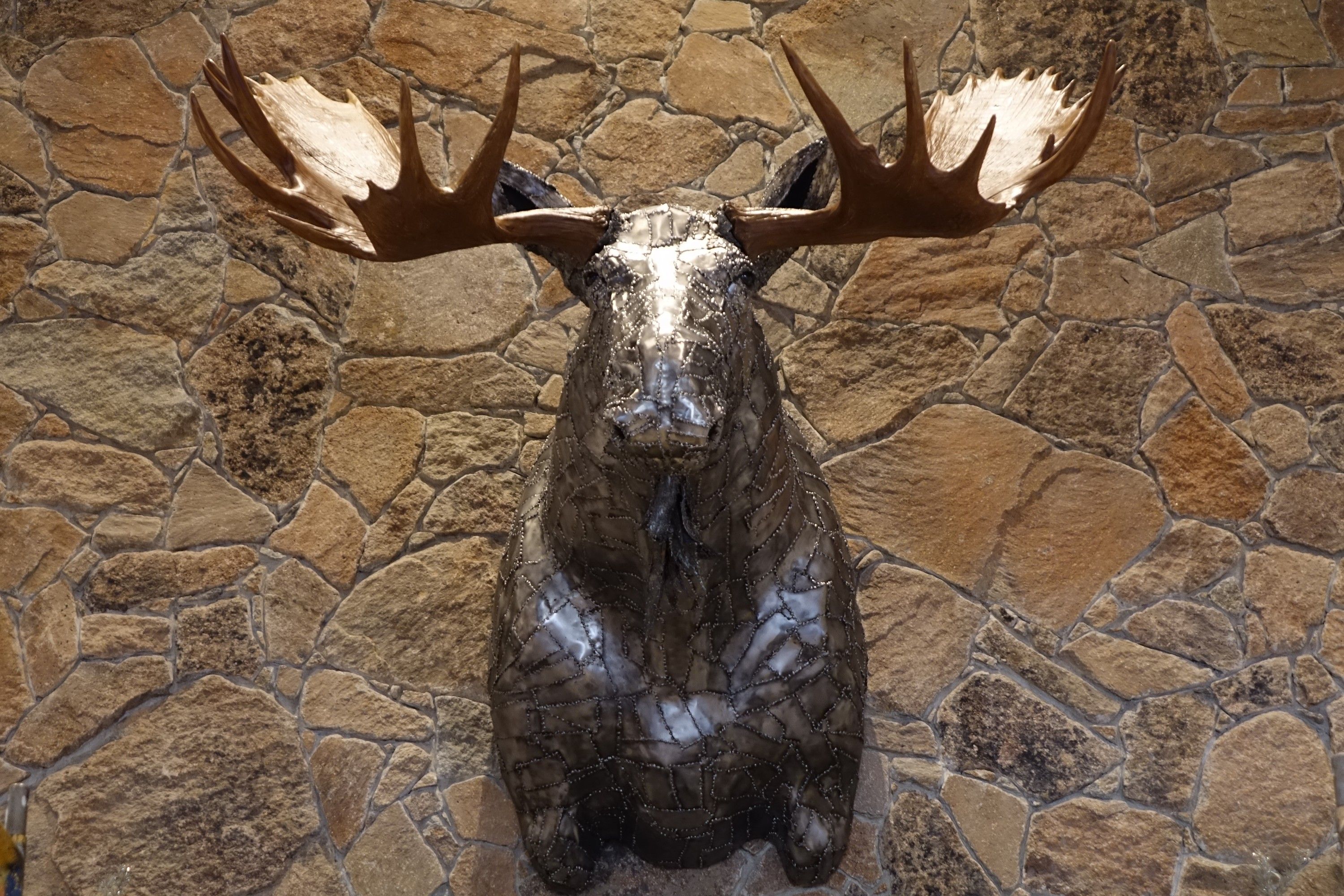 A welded steel moose head mounted on a stone wall, facing the camera, with broad reproduction antlers to either side