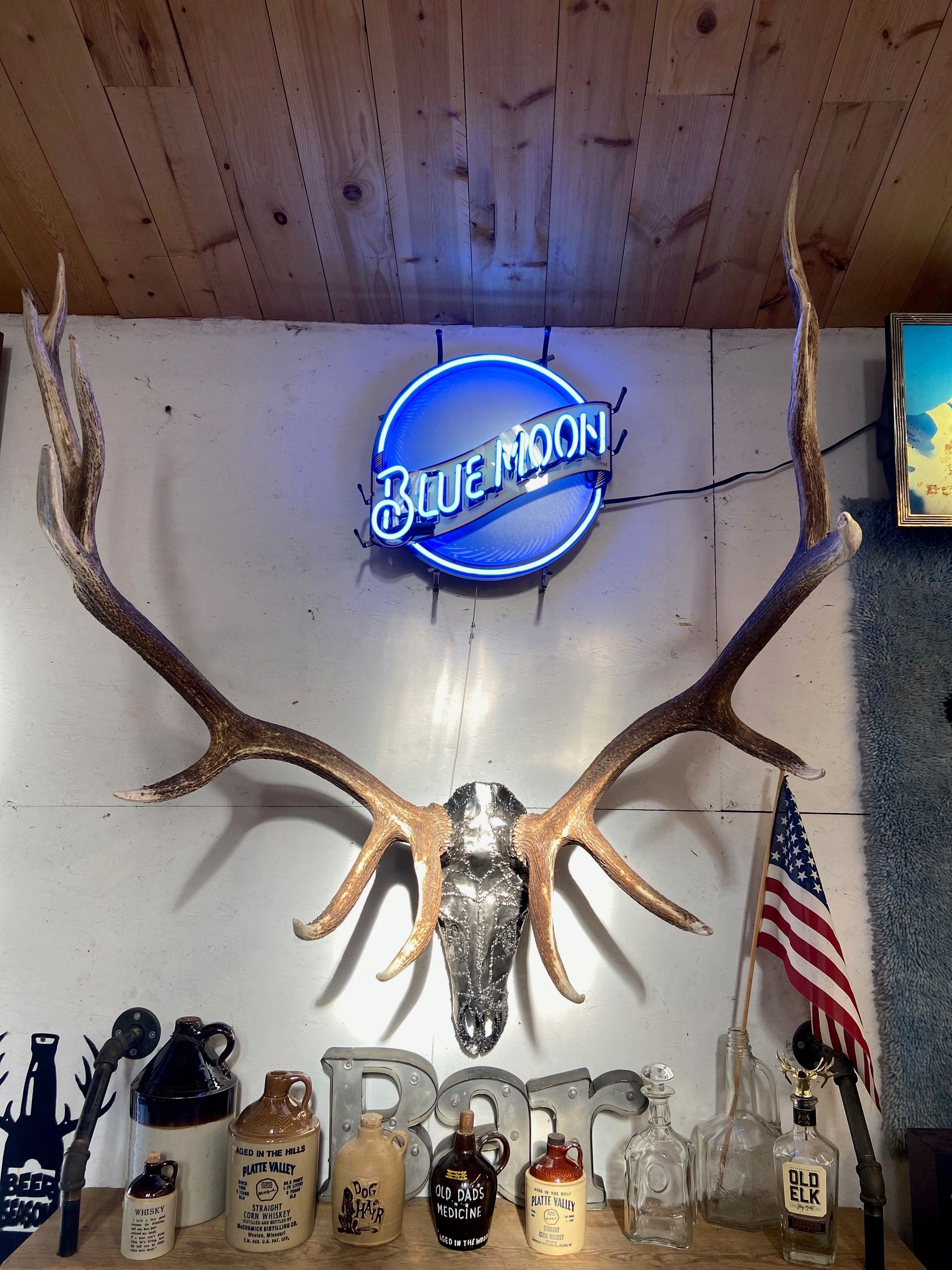 A steel-clad elk skull with natural horns mounted on a white plank wall, a neon sign glowing above it and a shelf of bottles below