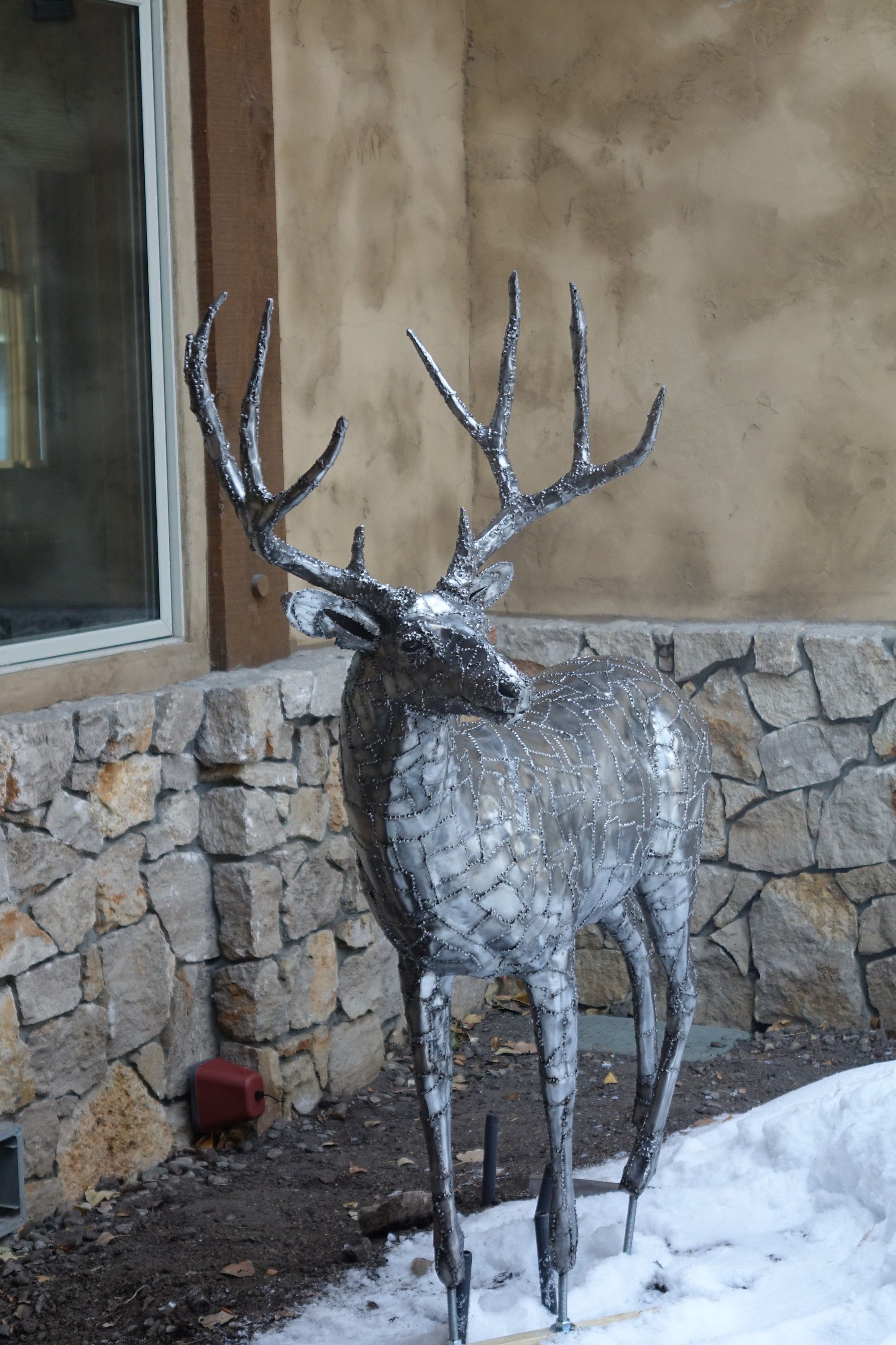 A life-size steel mule deer buck standing on gravel beside a stone house wall, its antlers spread above its head