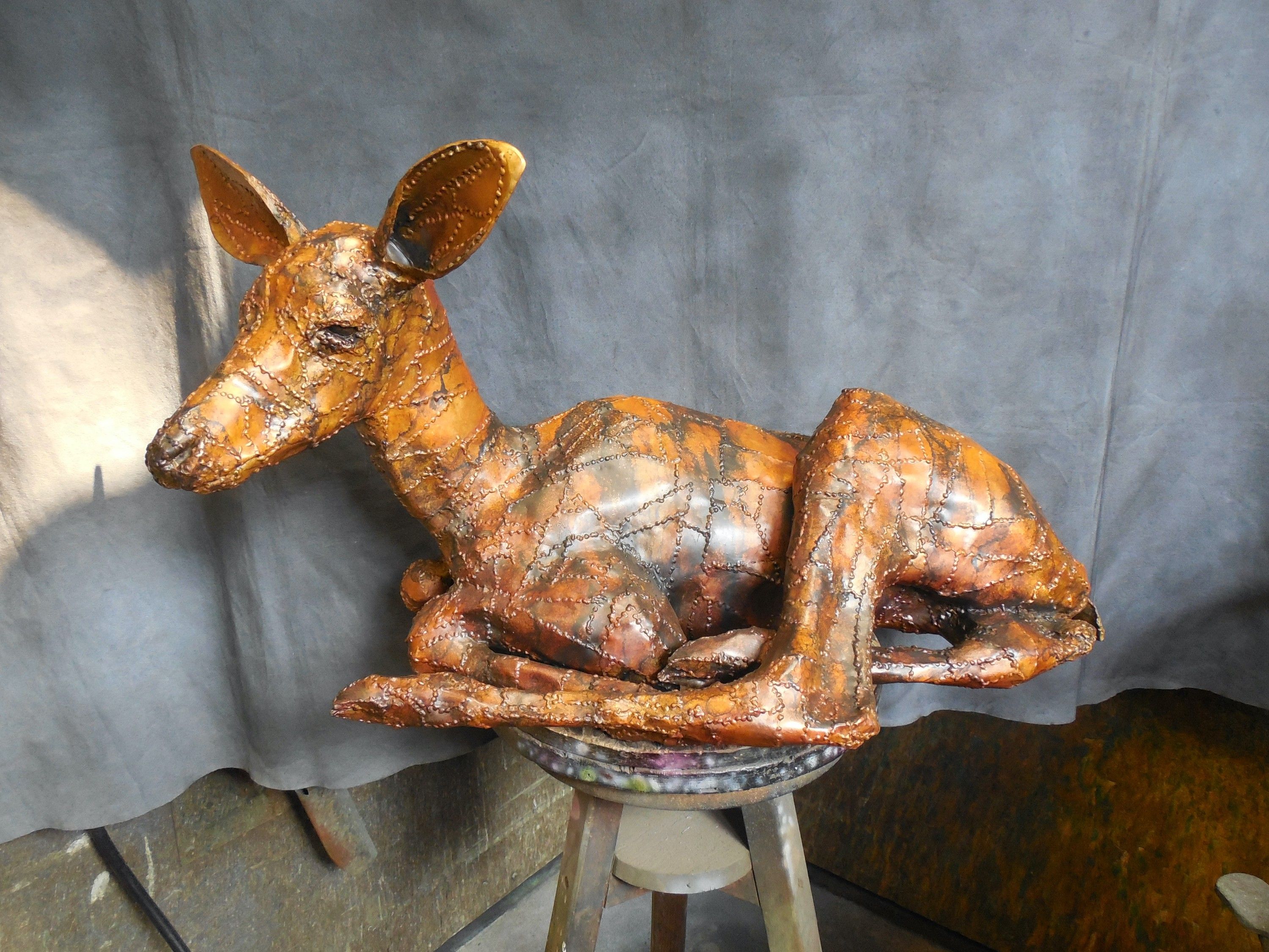 A bronze elk calf lying with its legs folded beneath it and its head raised, on a stool against a grey studio backdrop