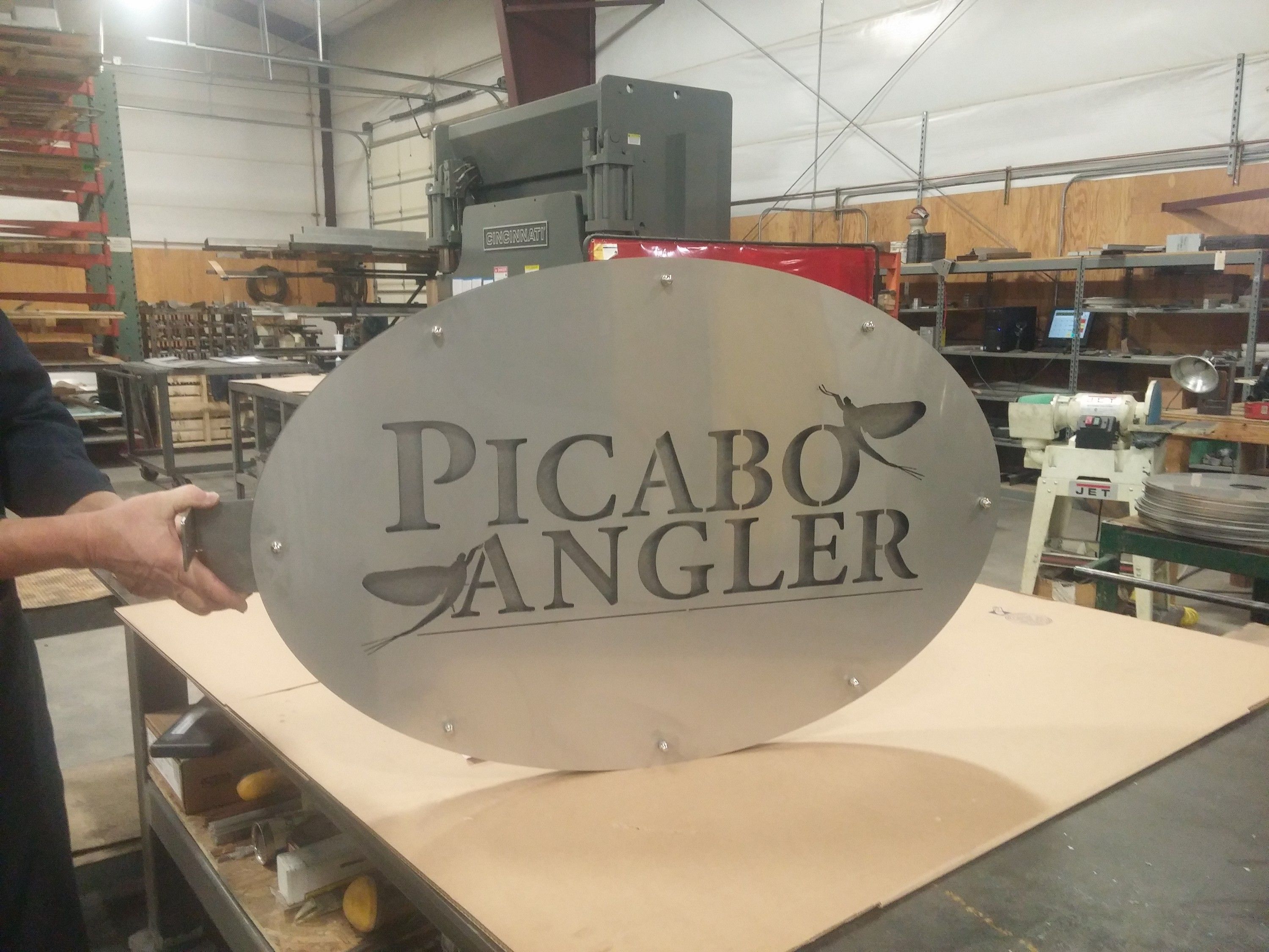 The oval Picabo Angler sign with mayfly cutouts, held upright in the workshop