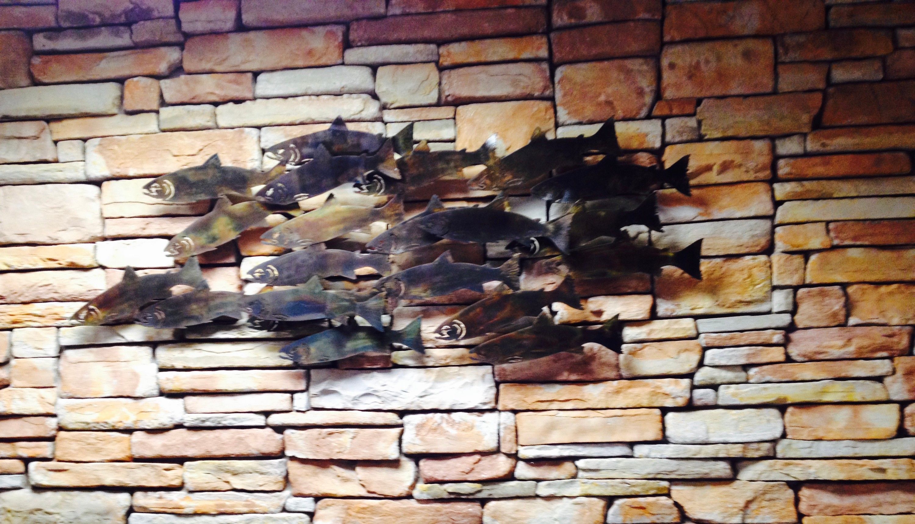 A school of patinaed steel fish mounted across a dry stone wall, angled as though moving together through the stonework