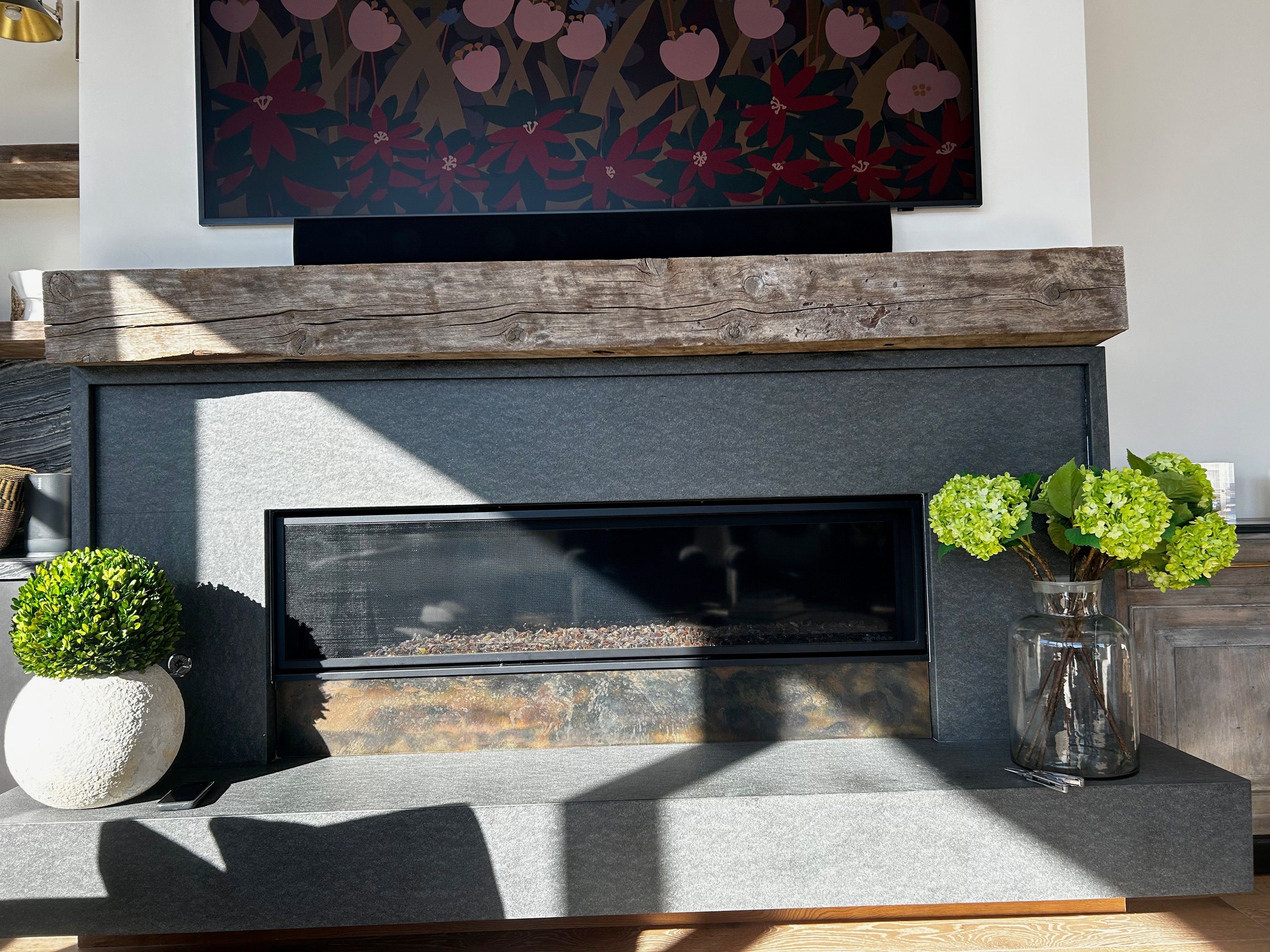 A long horizontal fireplace in a dark grey stone surround below a rough timber mantel, a mottled patina steel panel running the full width of the opening beneath the firebox glass, with a glass jar of green hydrangeas on the hearth to the right