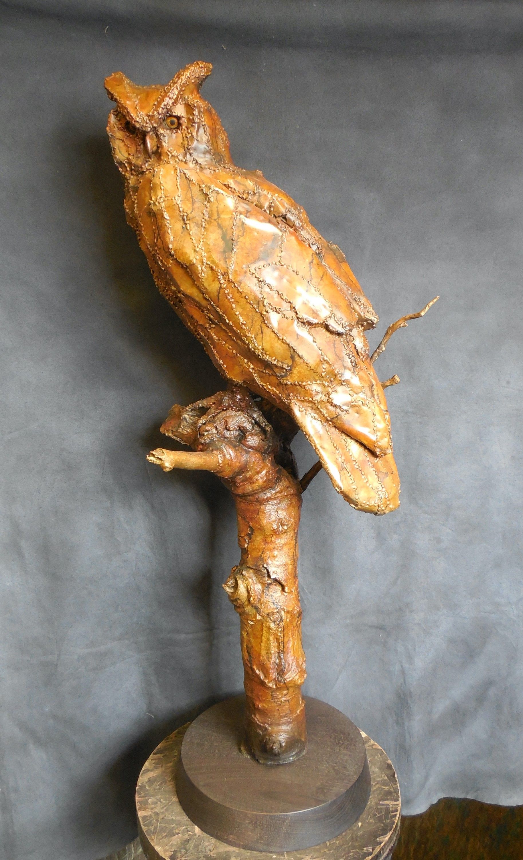 A bronze owl with raised ear tufts perched on a cut branch, seen from behind with its head turned, against a grey studio backdrop