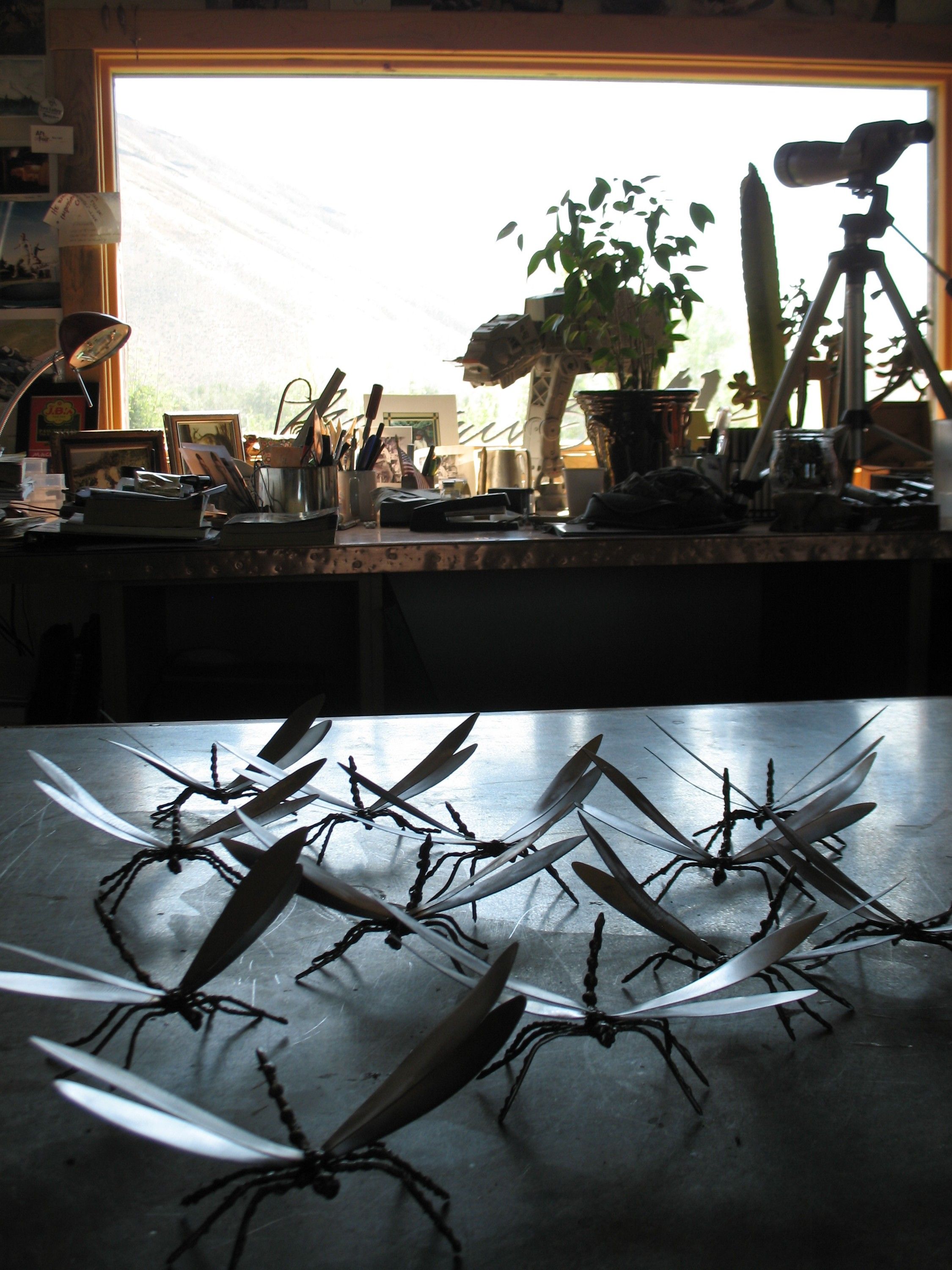 A dozen polished steel dragonflies standing on a steel bench in the studio, backlit by a wide window with a hillside beyond