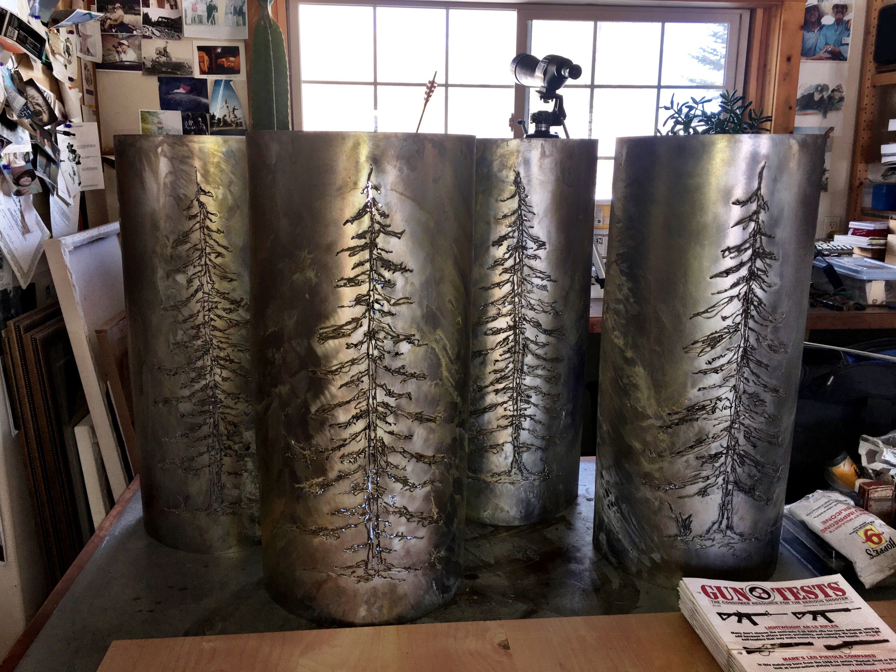 Four cylindrical patina steel sconces standing on a workbench, each cut through with the silhouette of a tall pine tree