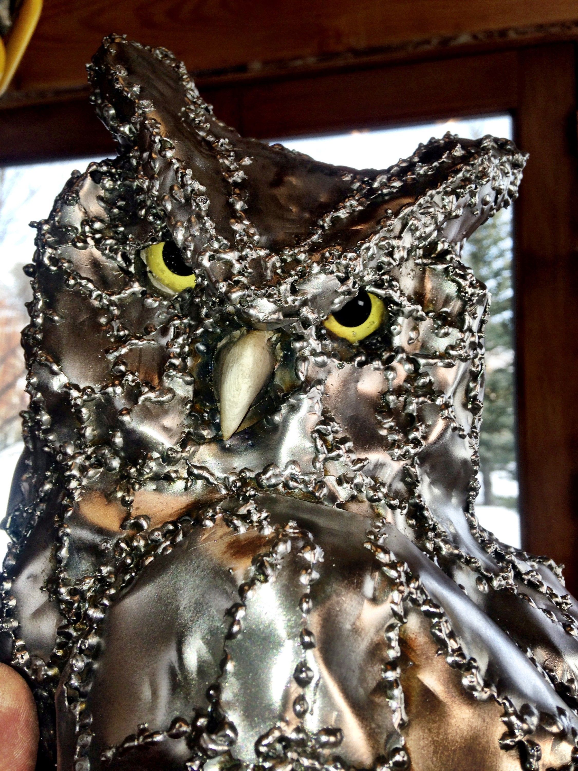 The steel owl's head filling the frame, its yellow glass eyes and pale beak set among overlapping steel plates, a snow-covered window behind