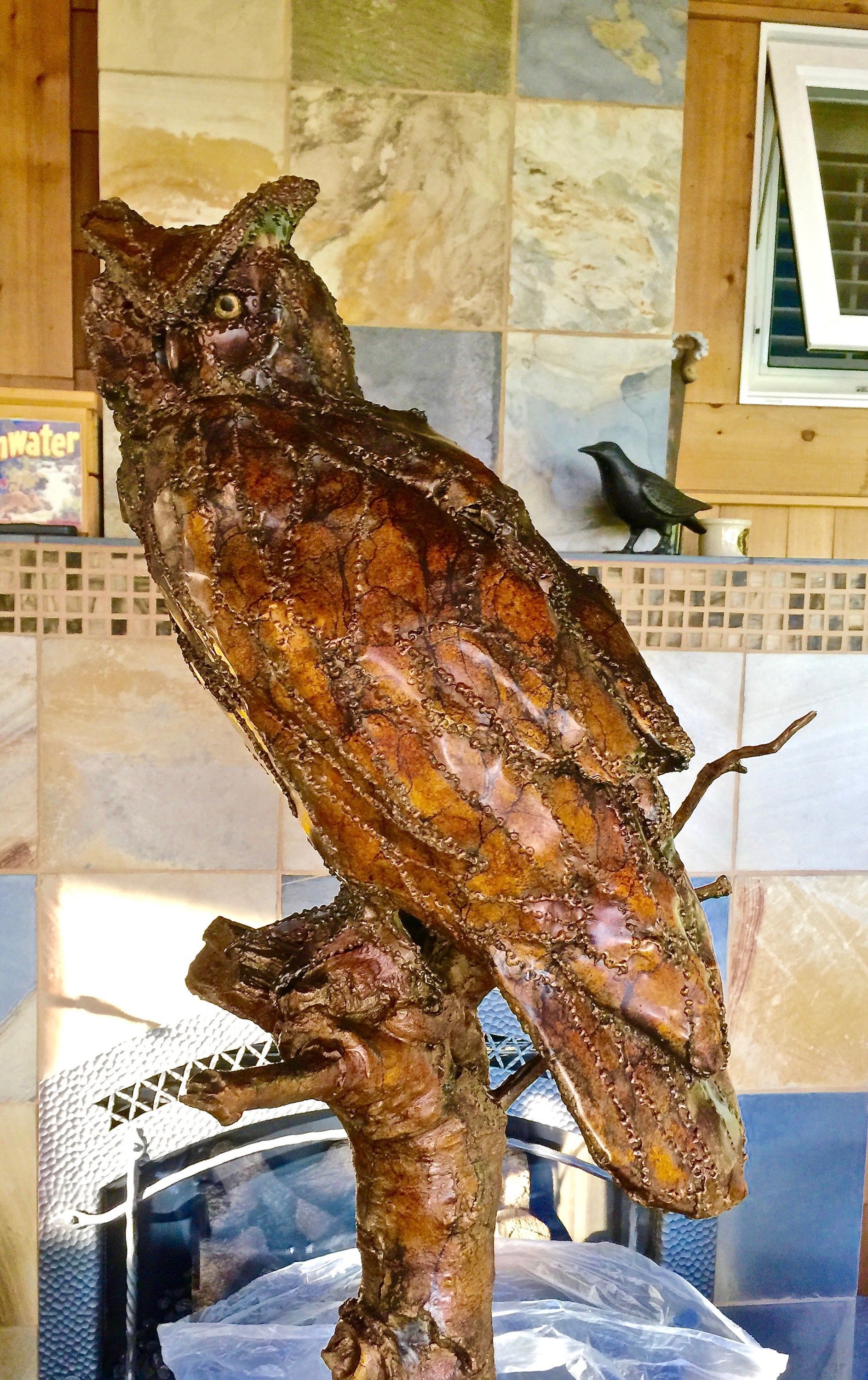 The bronze owl on its branch in front of a tiled fireplace wall, a small dark bird sculpture on the ledge behind it