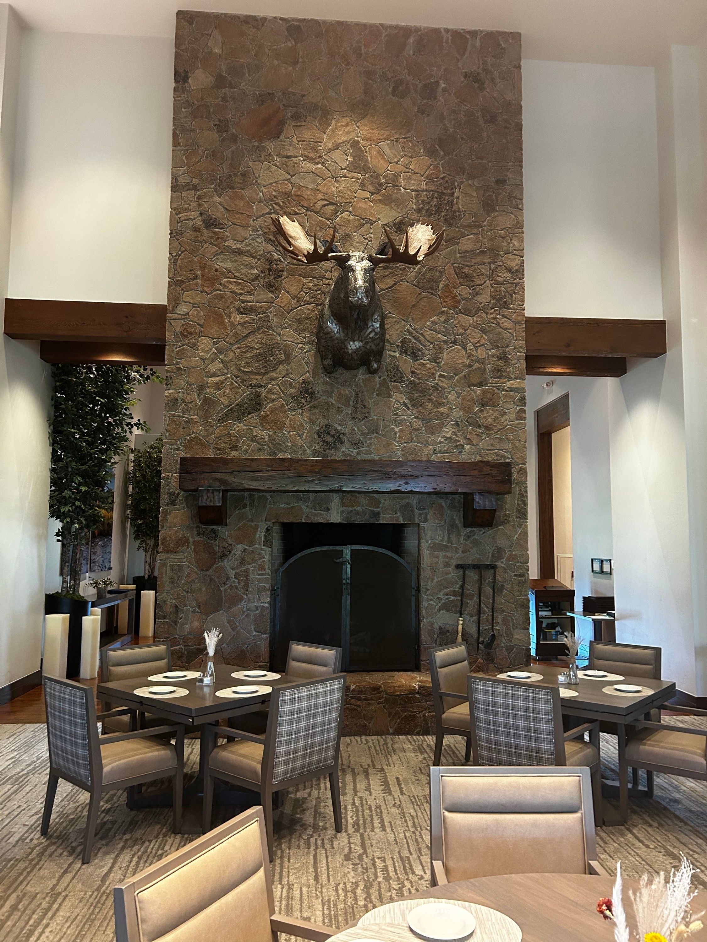 The moose head seen across a set dining room, small on the full height of the stone chimney above the fireplace