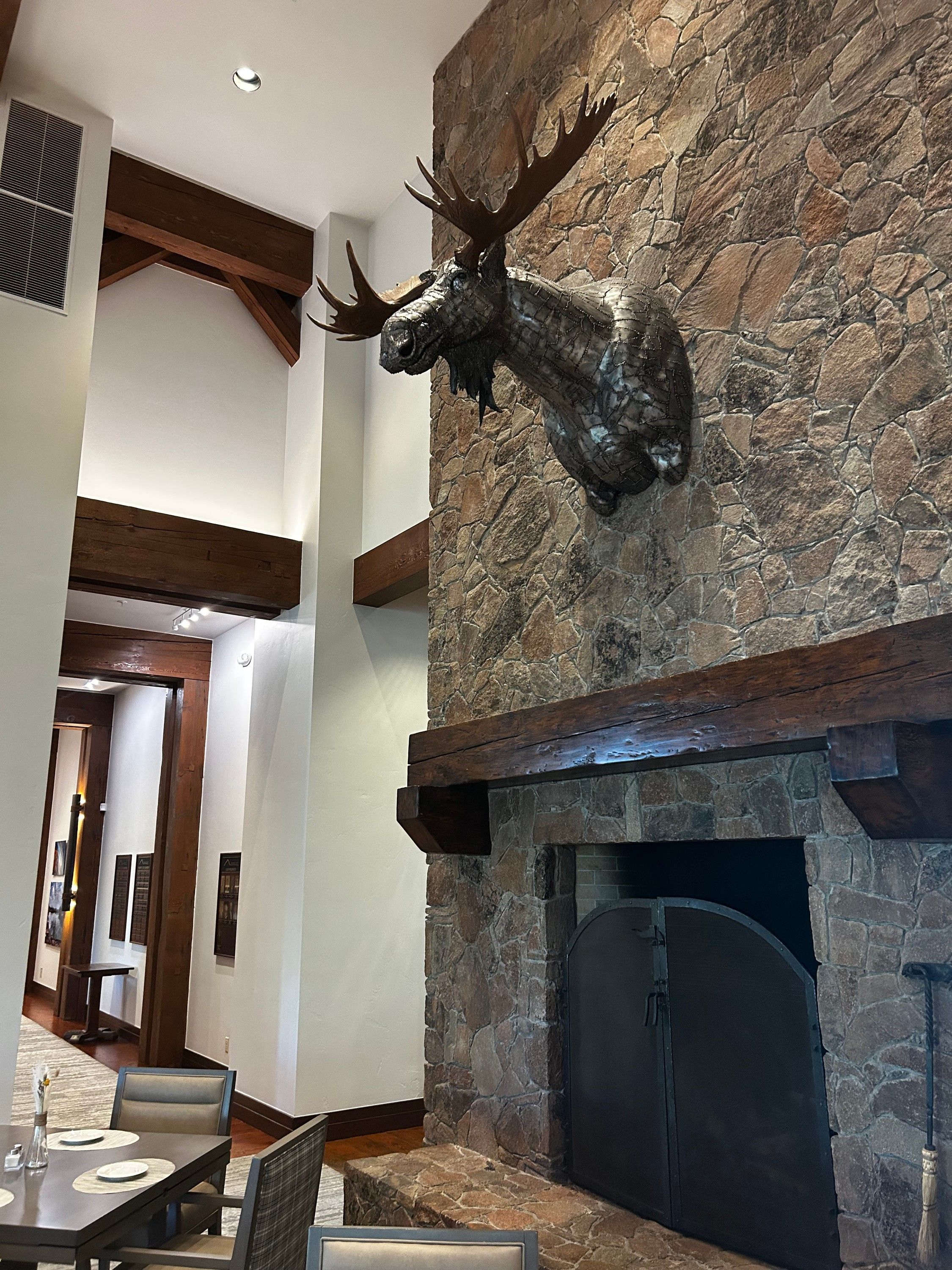 The moose head from the side, high on the stone chimney above the fireplace's iron doors, with a set dining table in the foreground and a lit corridor beyond