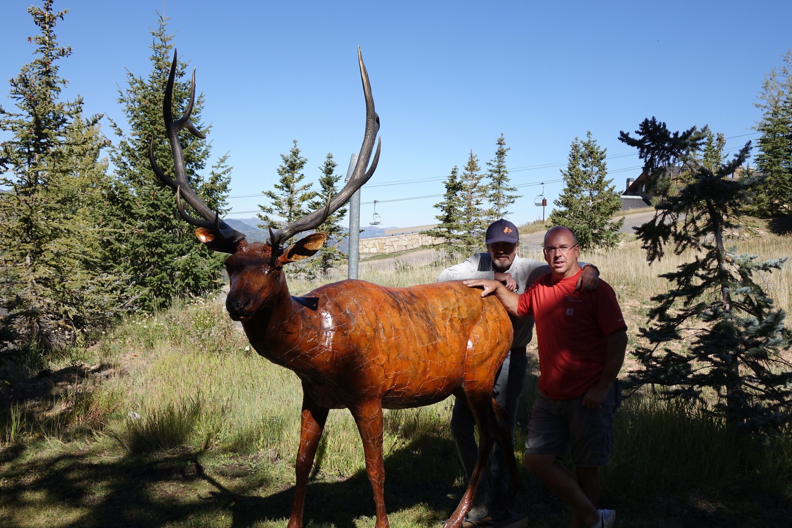 Two men standing beside the bronze bull elk on a grassy slope below a ski lift