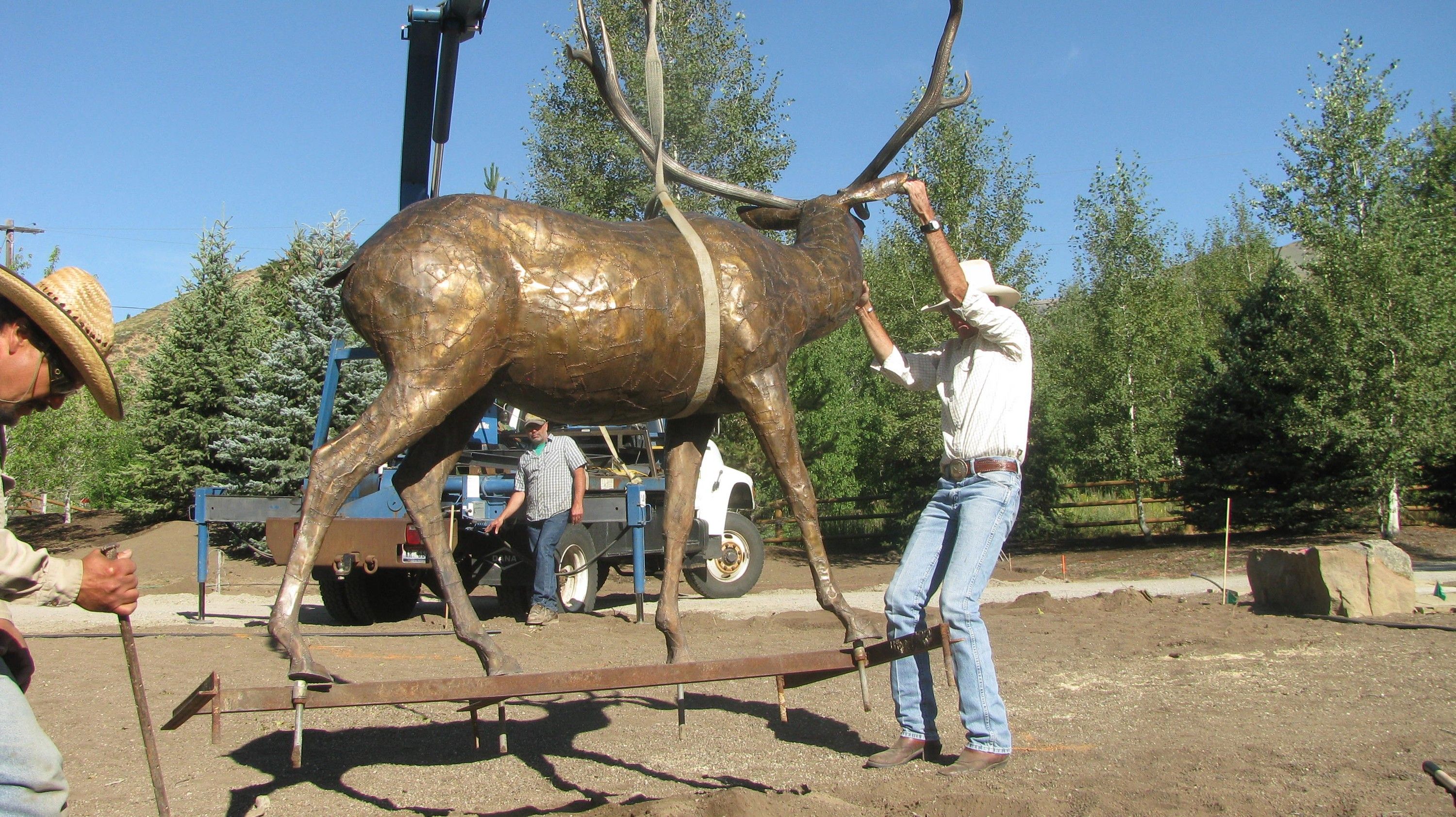 The bronze bull elk hanging in a crane strap, guided by hand onto the steel frame of its base